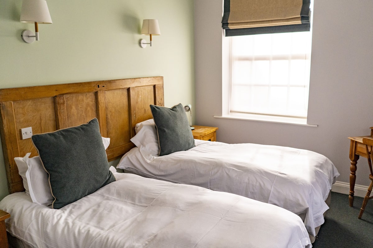 Una habitación con dos camas individuales, cabeceros de madera, ropa de cama blanca, dos mesitas de noche y una ventana con vistas.