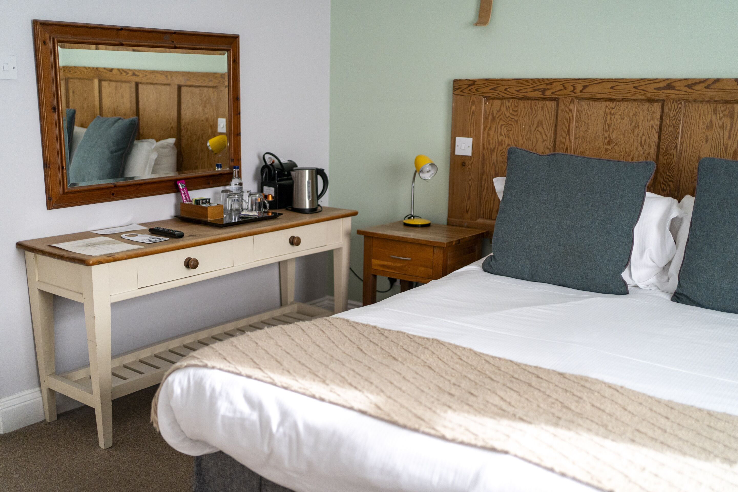 A hotel room with a wooden headboard, a large bed with white bedding, a wooden nightstand with a lamp, a desk with a mirror, and a small cabinet.