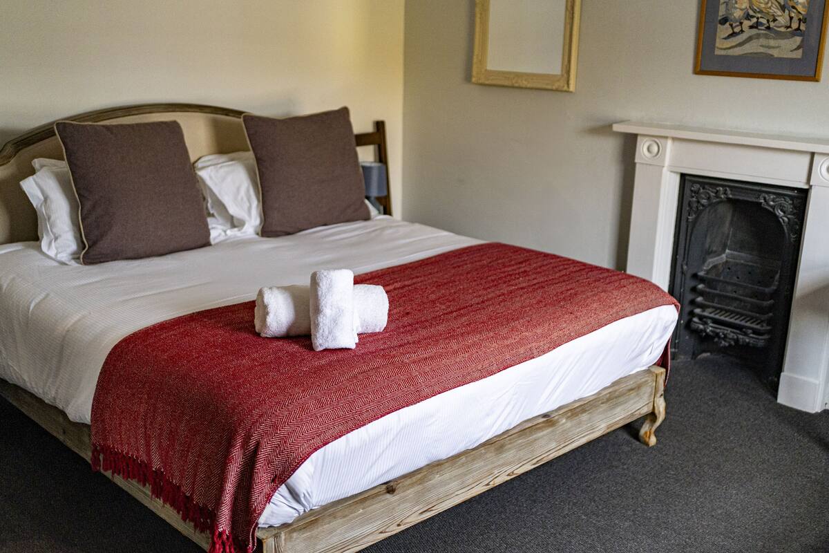 Una cama con estructura de madera, ropa de cama blanca y una colcha roja. Hay dos almohadas, una marrón y una blanca. Una toalla blanca está colocada sobre la cama.