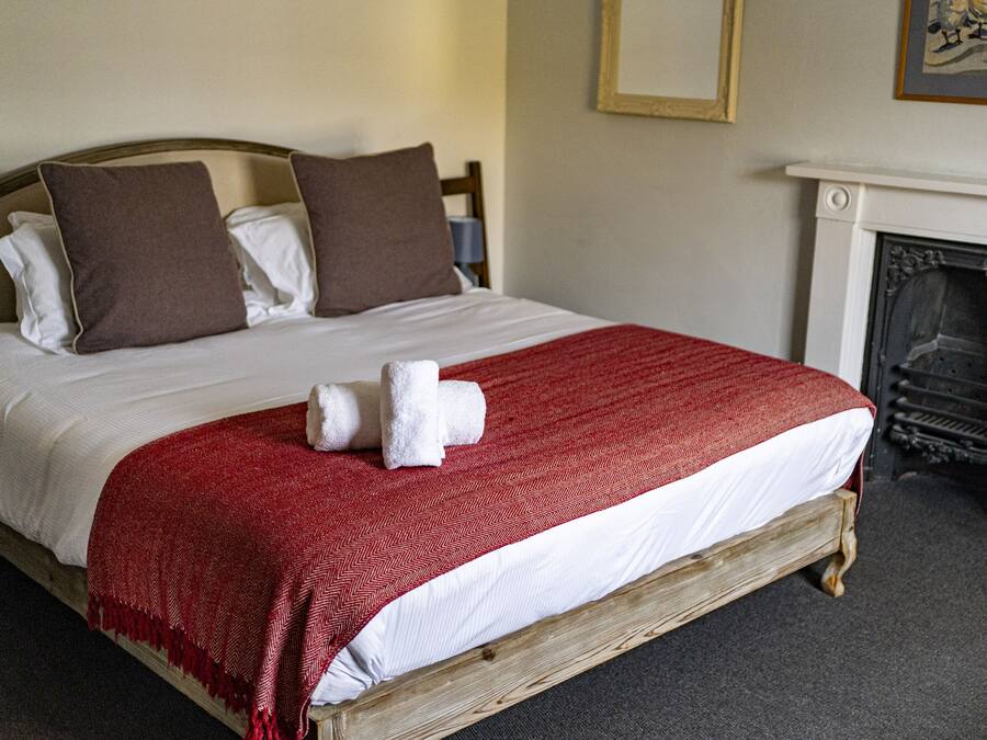 Una cama con estructura de madera, ropa de cama blanca y una colcha roja. Hay dos almohadas, una marrón y una blanca. Una toalla blanca está colocada sobre la cama.