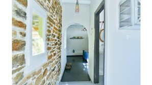 A narrow hallway with a stone wall on one side and a white wall on the other. There is a door on the left and a window on the right. - Amazing 1-Bedroom House in Tinos (Tinos)