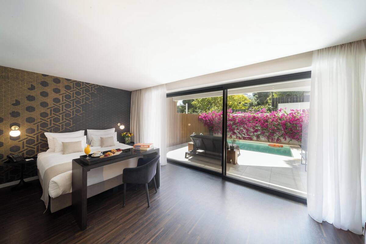 A hotel room with a bed, a desk with a laptop, a chair, and a view of a swimming pool and garden.