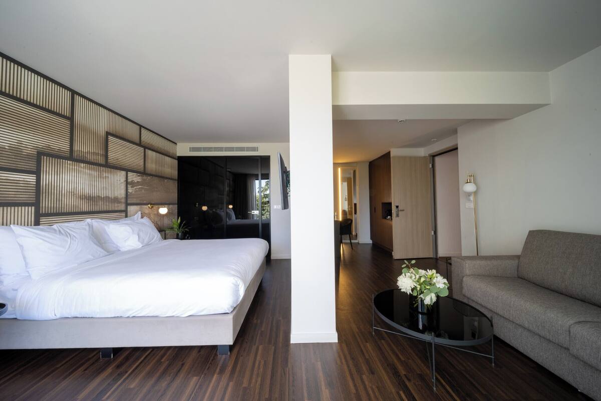 A modern hotel room with a large bed, a sofa, a glass coffee table, and a decorative vase.