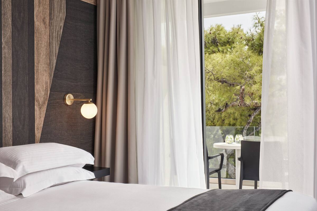 A modern hotel room with a large bed, a view of the outdoors, and a small table with chairs.