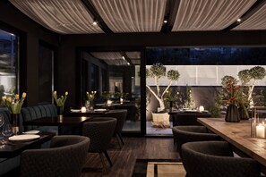 A modern dining area with dark wood furniture, candlelit tables, and a view of a garden through a glass wall. - Azur Hotel (Vari-Voula-Vouliagmeni)