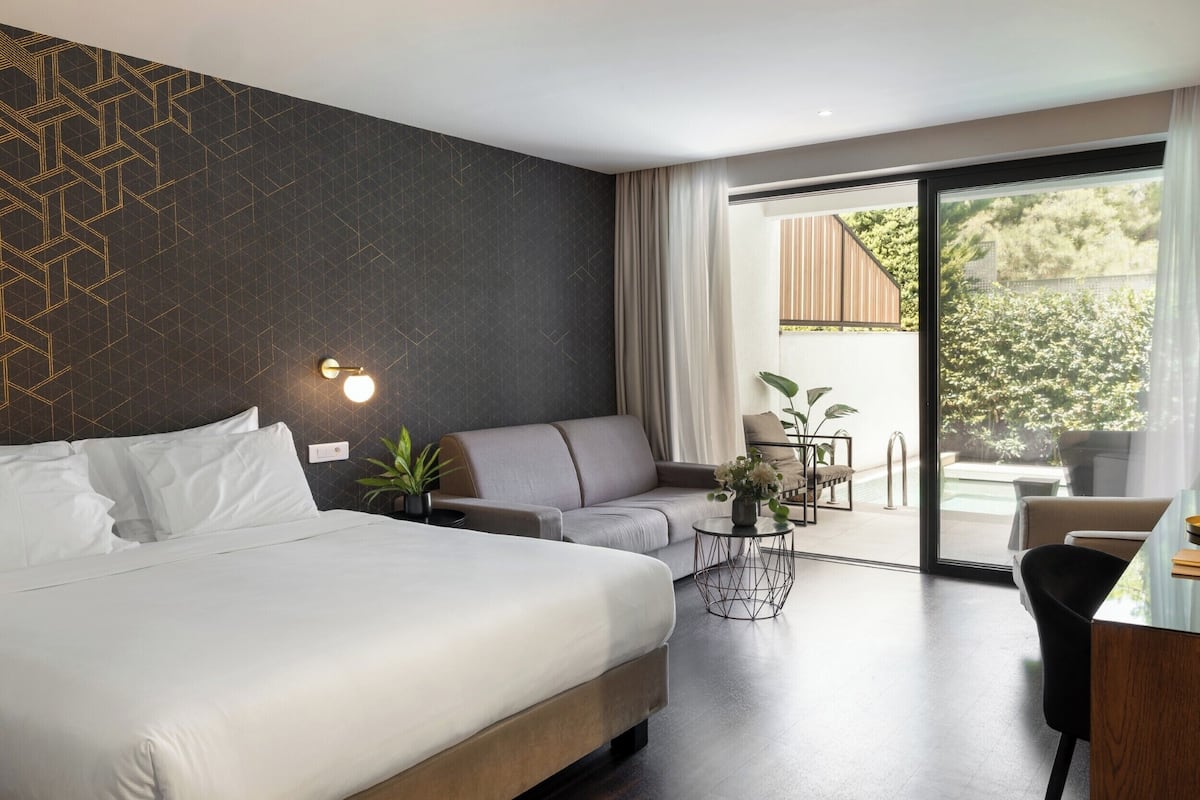 A modern hotel room with a large bed, a sofa, a small table with plants, and a view of a pool through a sliding glass door.