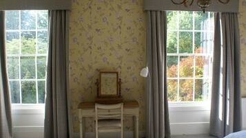 A room with a desk, chair, and a mirror. There are two windows with curtains, and a chandelier is visible.