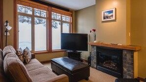 A living room with a sofa, ottoman, fireplace, TV, and a window with a view of snow-covered trees. - Ski in Ski out condo with private Hot Tub backing on Golf course. 2 bed 2 bath. (Sun Peaks)