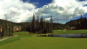 A golf course with a pond, surrounded by tall trees and a few buildings. - Ski in Ski out condo with private Hot Tub backing on Golf course. 2 bed 2 bath. (Sun Peaks)