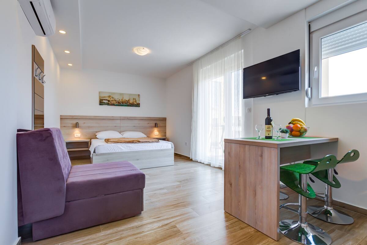A modern hotel room with a bed, a sofa, a small kitchenette, and a TV.