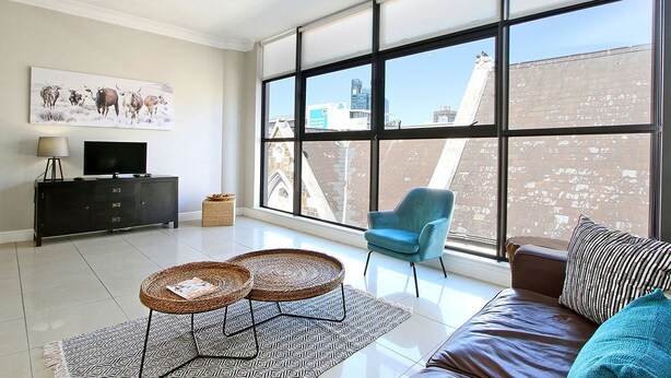 A modern living room with a large window, a sofa, a blue armchair, a television on a black cabinet, and a decorative wall piece. - Hill House 1 Bedroom (Cape Town)
