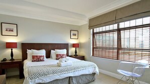 A neatly arranged bedroom with a large bed, two bedside tables with lamps, a clear plastic chair, and a window with blinds. - Hill House 1 Bedroom (Cape Town)
