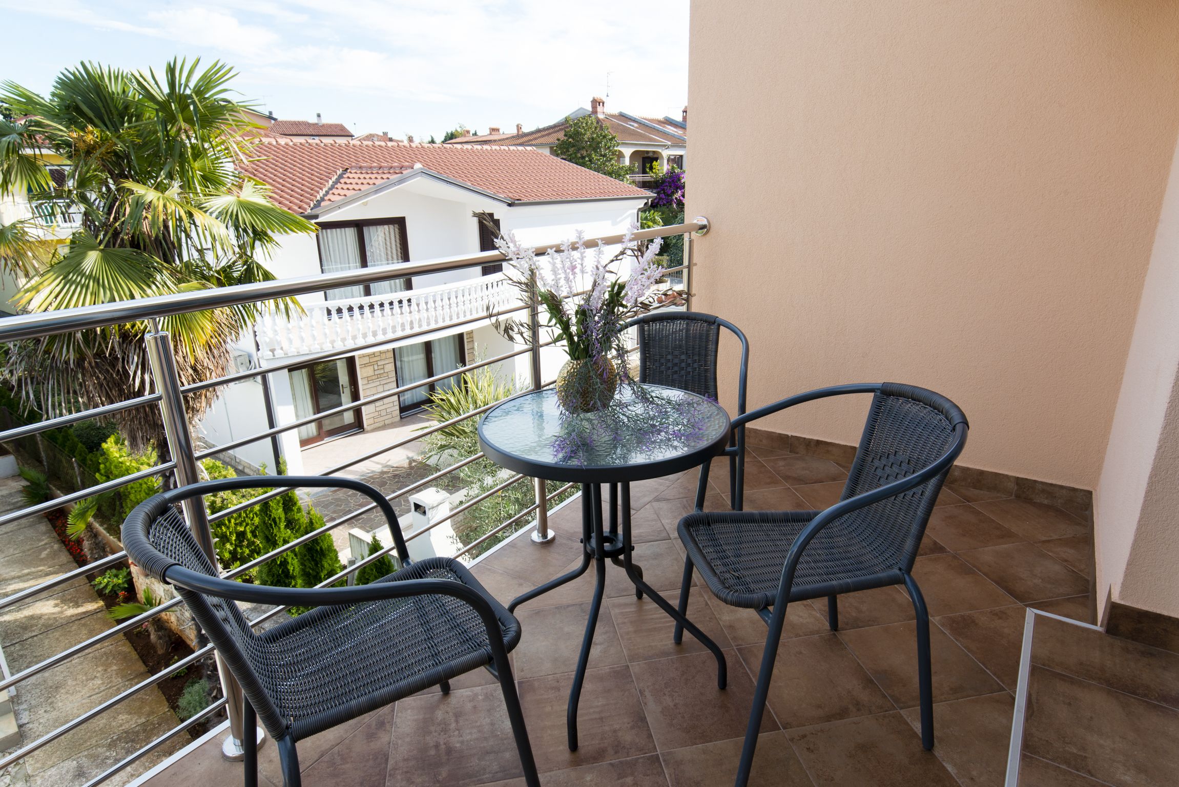 Een balkon met een ronde tafel en twee stoelen, met uitzicht op een tuin en naburige gebouwen.