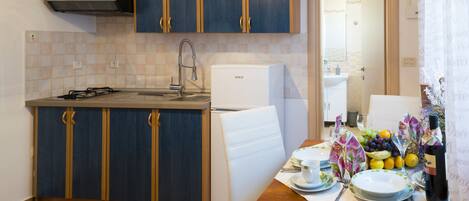A compact kitchen with a sink, stove, and white refrigerator. There's a dining table with plates, a bottle of wine, and a fruit bowl.