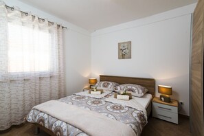 A bedroom with a bed, two bedside tables, a nightstand, a window with curtains, and a wall-mounted artwork.