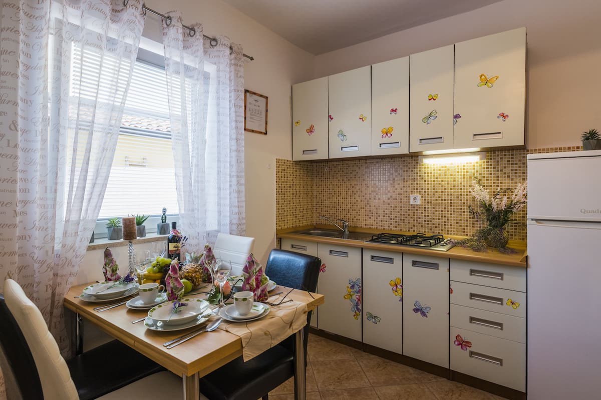 A kitchen with a dining table set for two, a window with sheer curtains, and cabinets with butterfly decals.