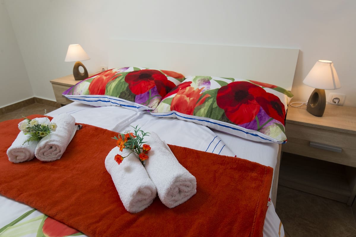 A neatly made bed with floral-patterned pillows, two rolled towels with flowers, and bedside lamps on wooden nightstands.