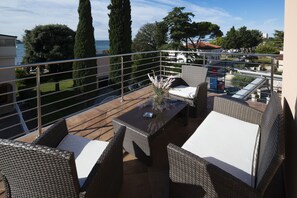 A balcony with wicker furniture, a table with a vase, and a view of the sea and trees.