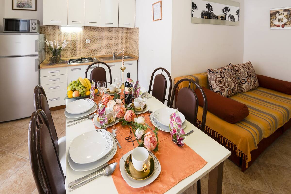 A dining area with a table set for four, including plates, cutlery, and a vase of flowers. A kitchen is visible in the background with a microwave and cabinets.