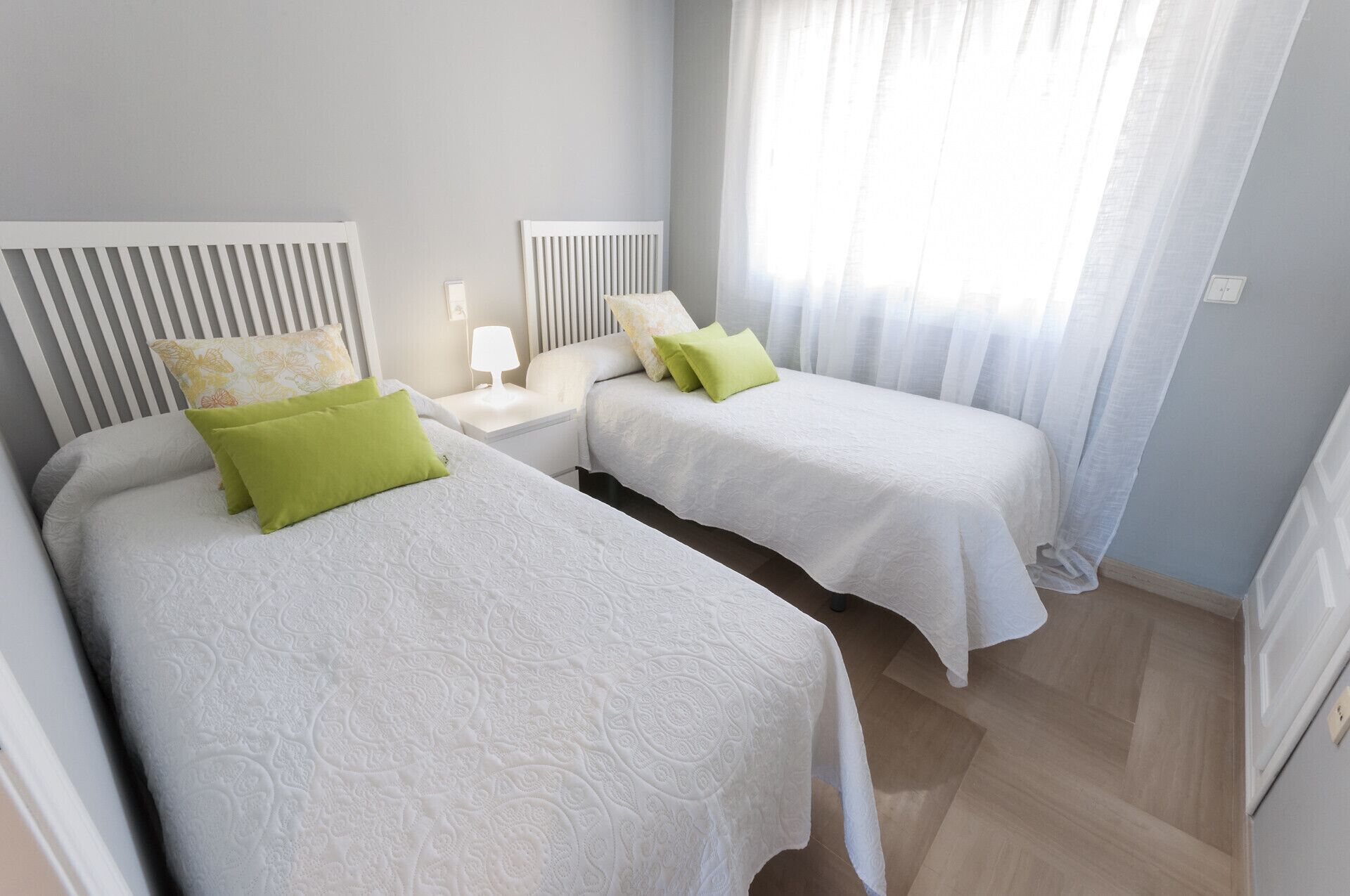 Et hotelværelse med to senge, et natbord med lampe og et vindue med let gardin.