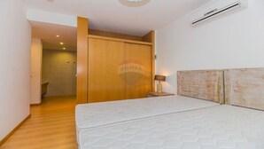 Un dormitorio con un armario de madera, una cama con colchón blanco, una lámpara de noche y un equipo de aire acondicionado en la pared.