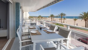 Un balcón con una mesa blanca lista para comer, con vista a una playa con palmeras y un cielo despejado.