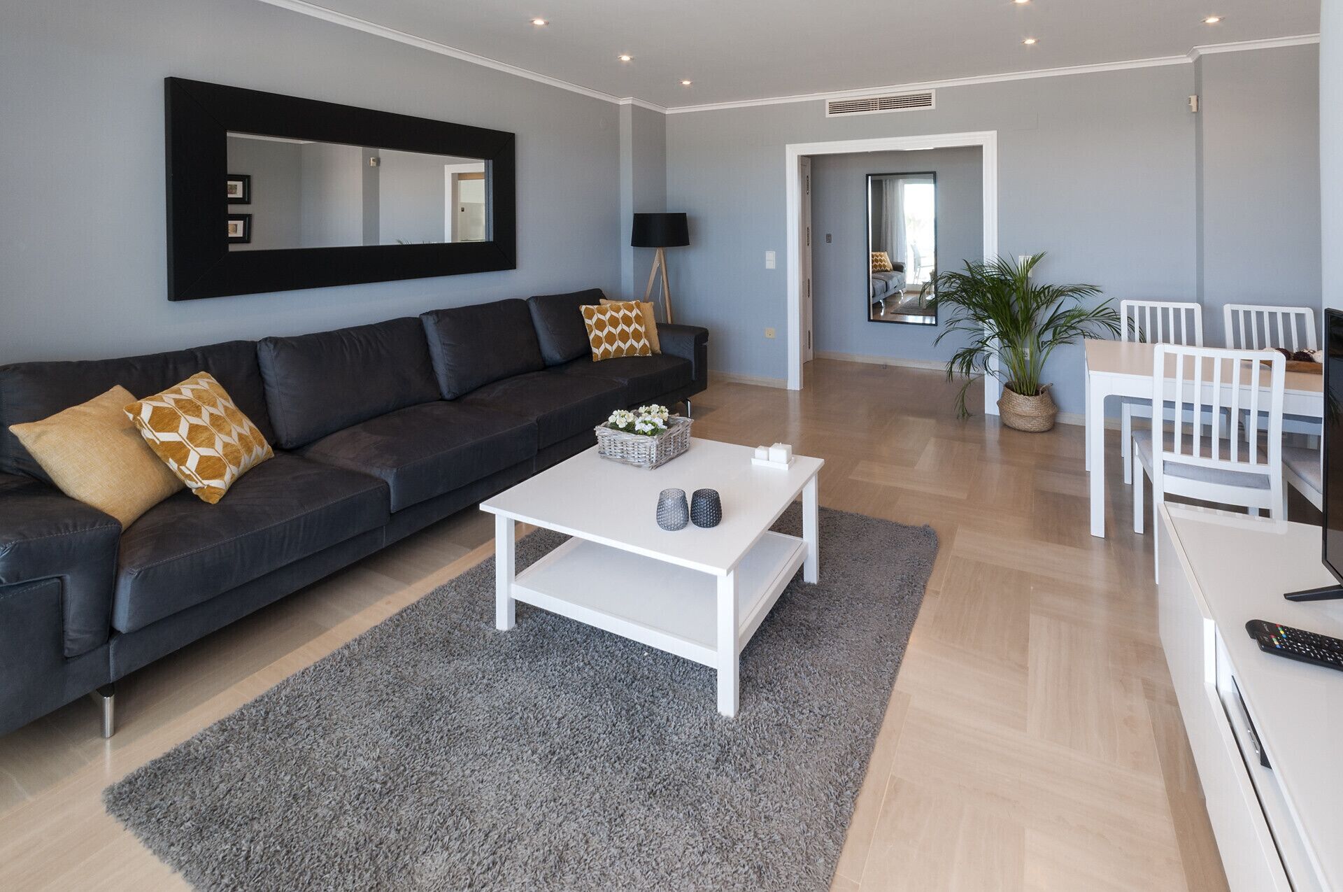 Een moderne woonkamer met een donkergrijze bank, een witte salontafel en een grijze vloerkleed. Er is een wit eethoek met een tafel en stoelen.