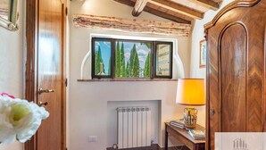 Een kamer met een houten plafond, een raam met uitzicht op bomen, een radiator en een lamp op een tafel.