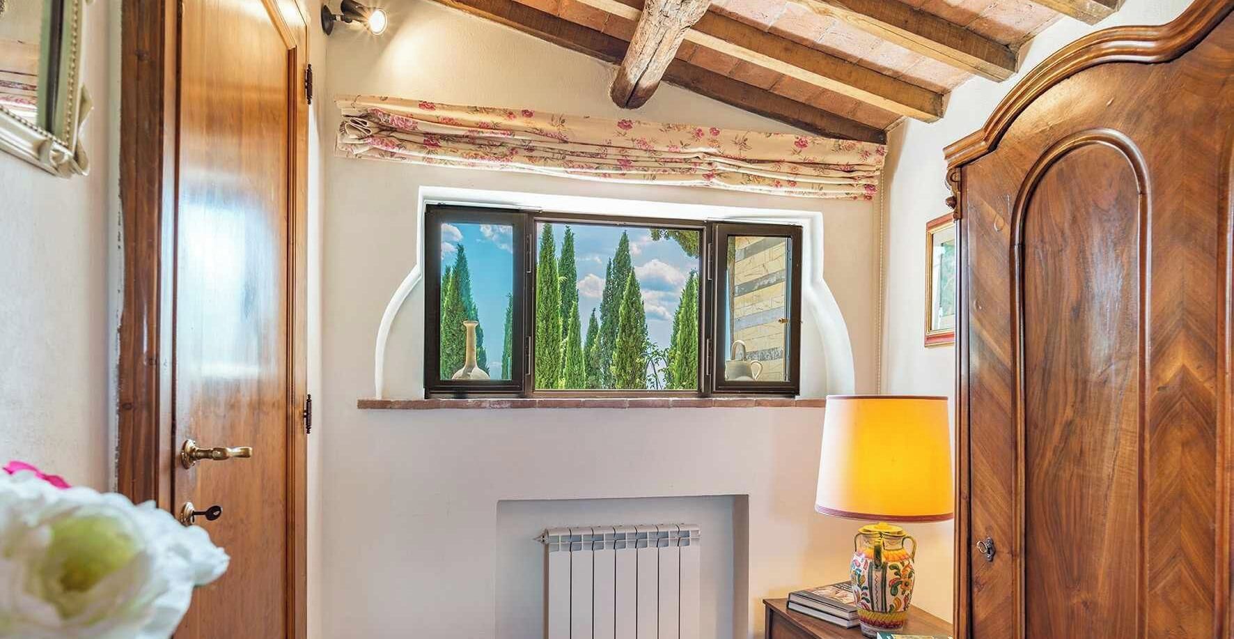 Ein Zimmer mit Holztür, Fenster mit Blick auf Bäume, Heizkörper und Lampe auf einem Nachttisch.
