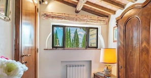 Een kamer met een houten deur, een raam met uitzicht op bomen, een radiator en een lamp op een bijzettafeltje.