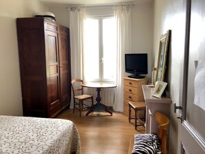 A room with a bed, a wooden wardrobe, a desk with a chair, a television, and a window with curtains.