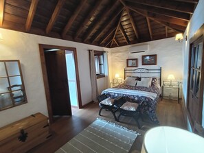 Une chambre avec un plafond en bois, un lit recouvert d’une literie à motifs floraux, deux tables de chevet avec des lampes et une fenêtre avec des rideaux.