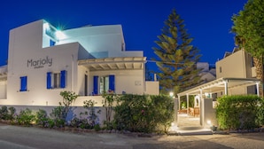 A modern building with the name "Marioly Studios" prominently displayed. It features a white facade with blue shutters, a covered outdoor seating area, and is surrounded by greenery. - Marioly Studios (Paros)