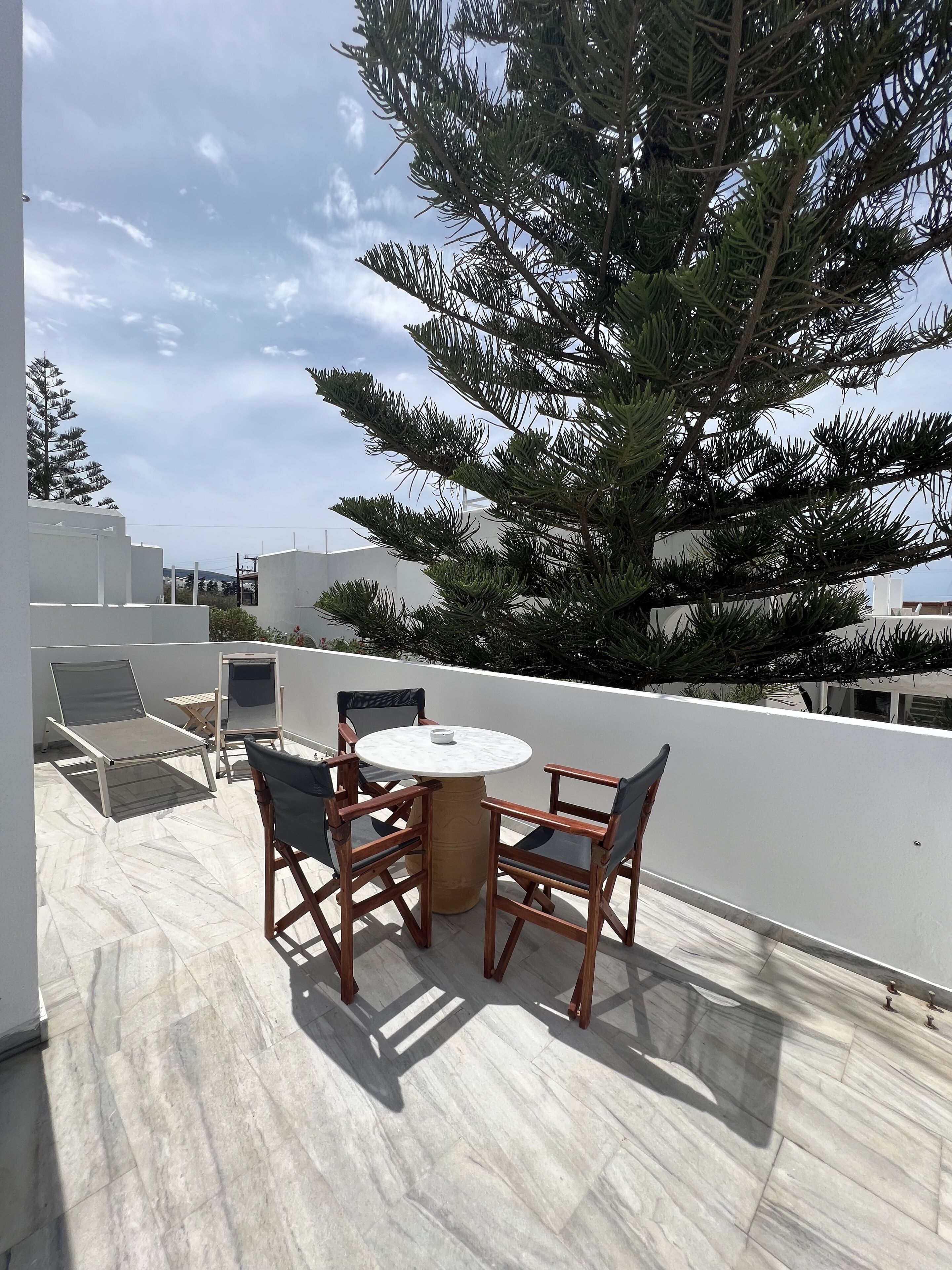 Una terraza en la azotea con una mesa redonda y cuatro sillas, rodeada por un muro blanco y un gran árbol.