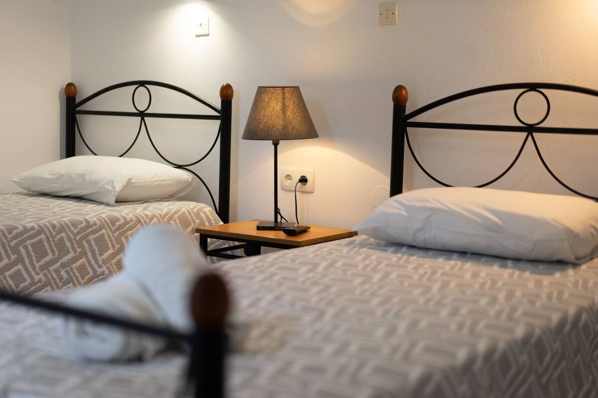 A hotel room with two single beds, each with a wooden headboard, a bedside table with a lamp, and a power outlet.
