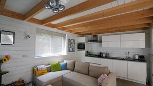 Un interior de cabaña acogedor, con techo de madera, kitchenette, sofá con cojines y ventana con persianas.