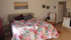 A bedroom with a bed, a chair, a nightstand, a picture on the wall, and a kitchen area.