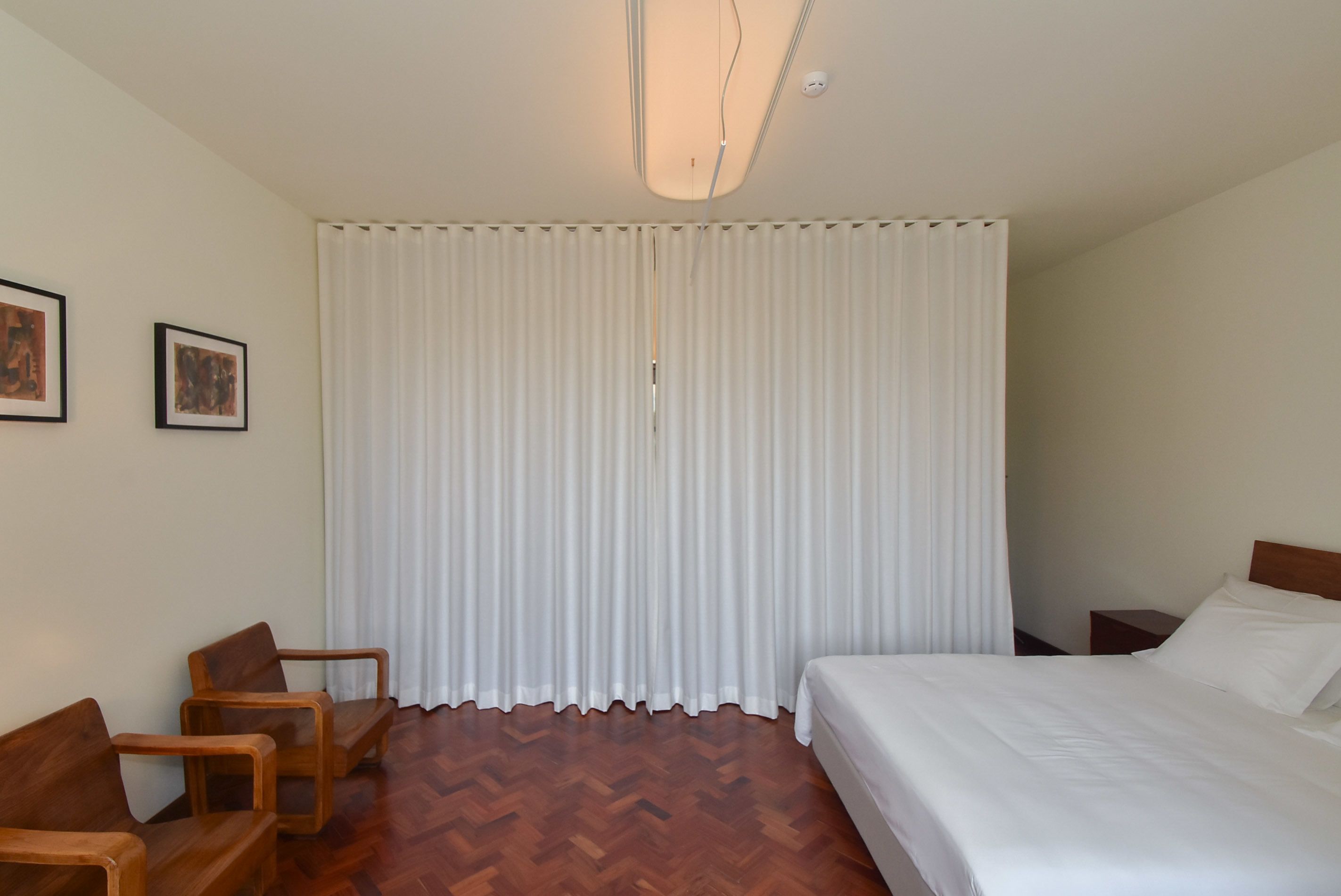 A hotel room with a bed, wooden chairs, a white curtain, and a ceiling light fixture.