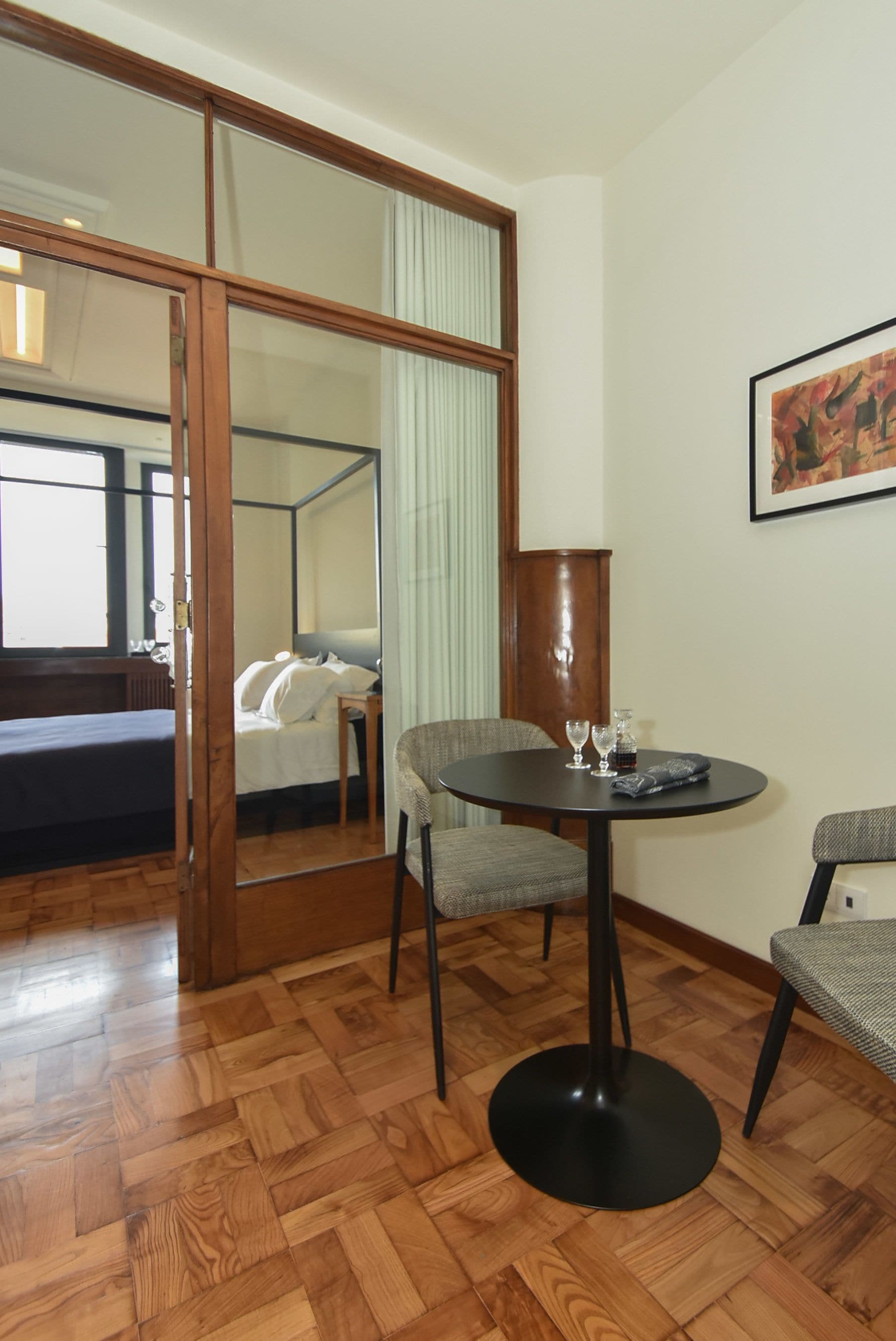 A hotel room with a wooden floor, a round table with chairs, a bed, a bedside table with a lamp, and a large mirror.