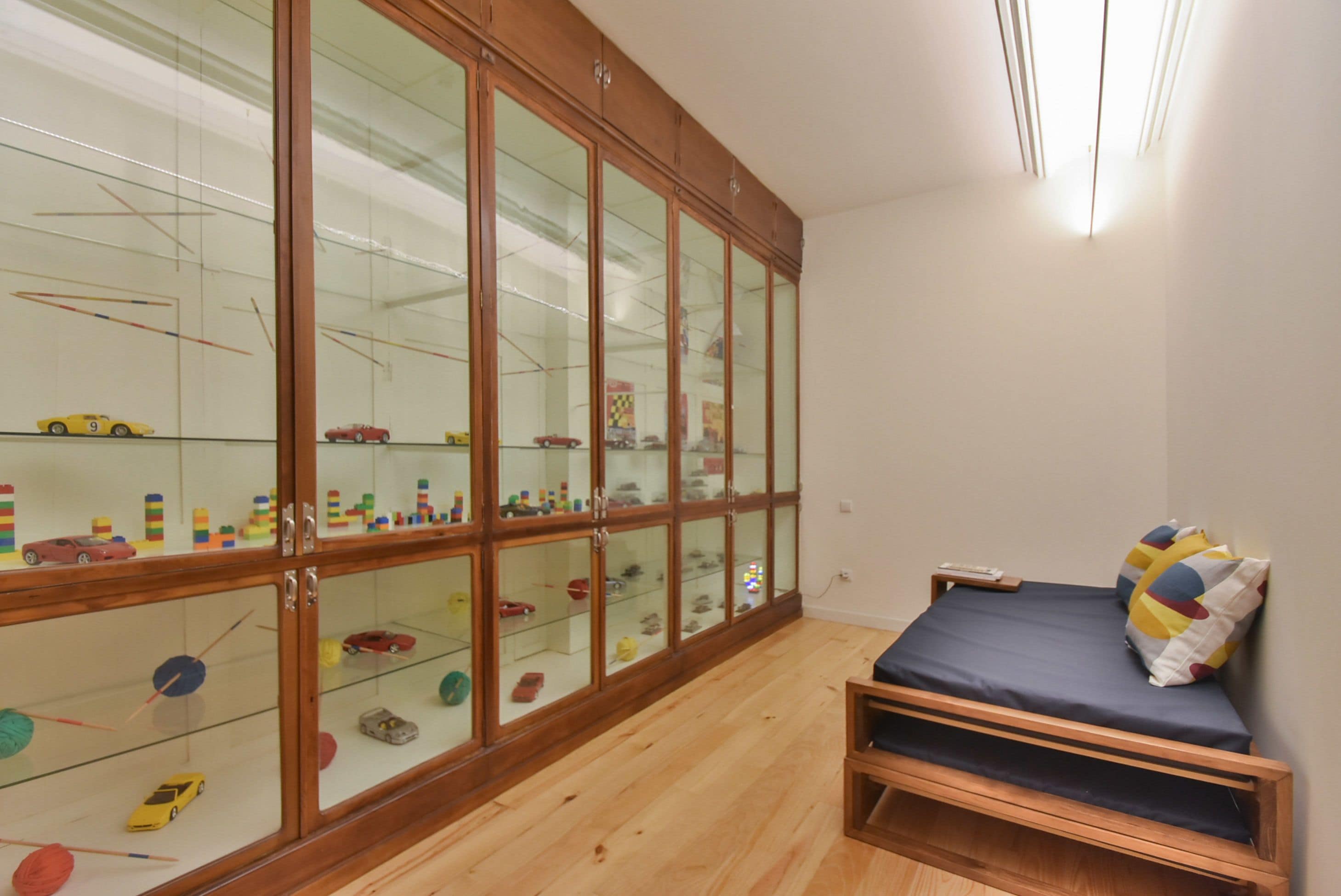 Une pièce avec une armoire en bois contenant des vitrines où sont exposés des jouets et des modèles. On y trouve un lit avec un matelas et des oreillers bleus, ainsi qu’une petite étagère en bois.