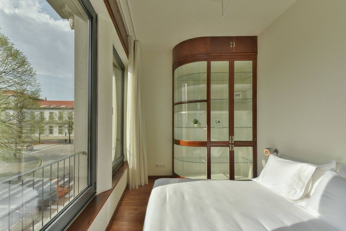 Un dormitorio con un ventanal grande, un armario de madera curvada y una cama con ropa de cama blanca.