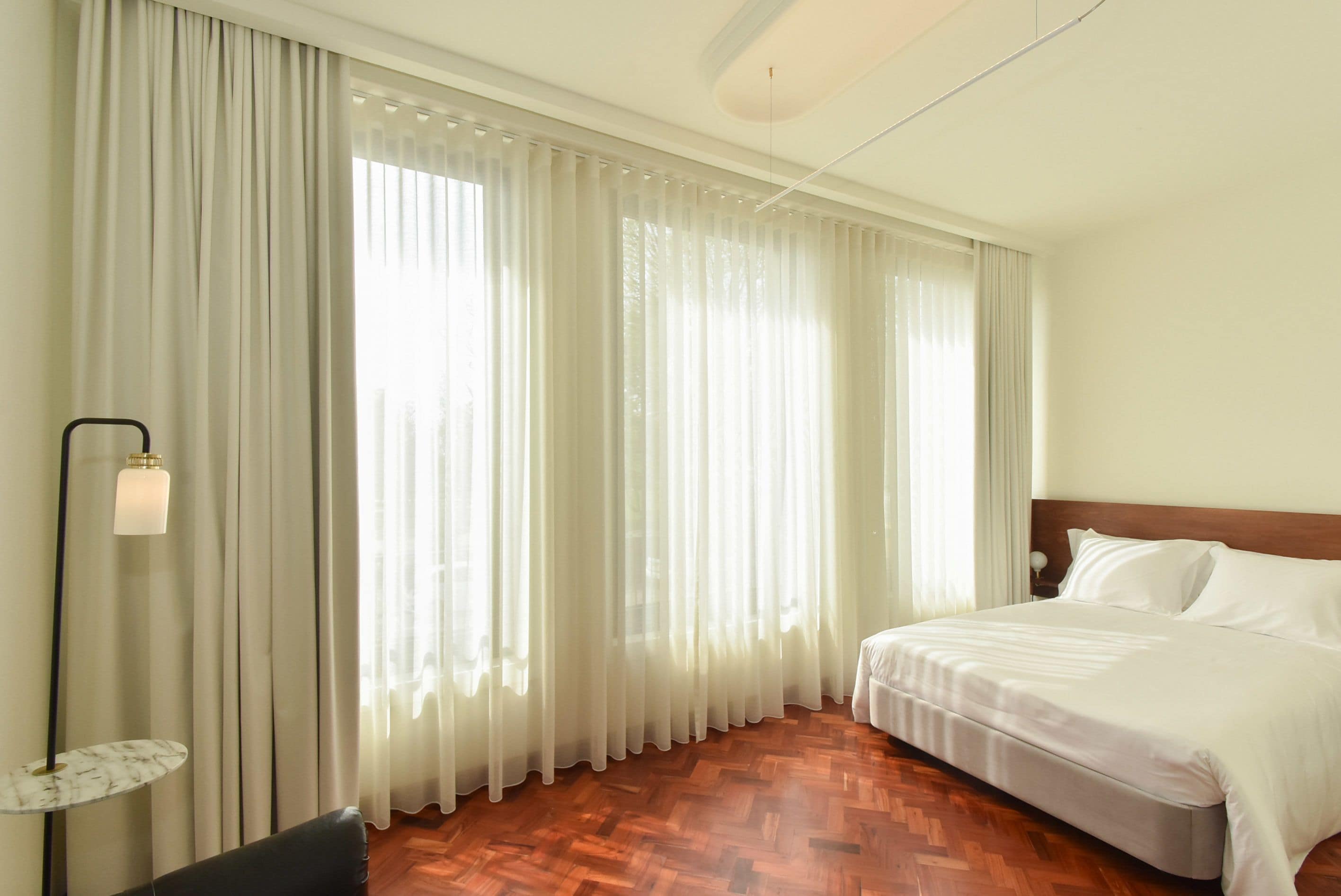 A hotel room with a bed, a nightstand, a lamp, and sheer curtains.