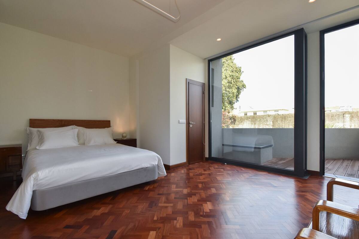 Un dormitorio con piso de madera en espiga, una cama con sábanas blancas, una mesita de noche, una puerta corrediza de vidrio a un balcón y vistas al exterior.