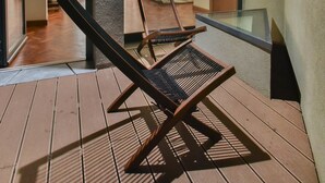 A wooden deck with two lounge chairs, a glass balustrade, and a view into a room with a bed and curtains. - República 157 (Porto)