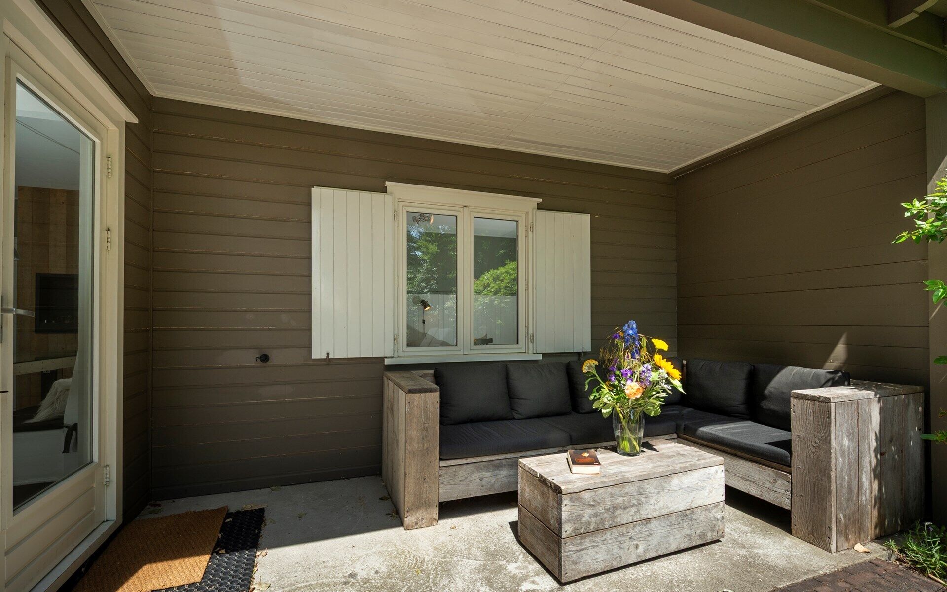 Un patio couvert avec un mur en bois foncé, une fenêtre à cadre blanc, un canapé noir, une table basse en bois et un vase rempli de fleurs.