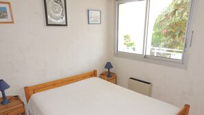 A bedroom with a wooden bed, a nightstand with a lamp, and a window with a view of trees. - Royan: T2 apartment near beach with parking (Royan)