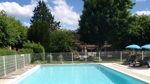 Una piscina rectangular con valla metálica, rodeada de árboles y un patio con sillas de descanso y sombrillas.