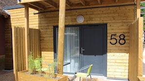 Un edificio de madera con el número '58' en la puerta, una jardinera de madera con plantas y una silla verde en el exterior.