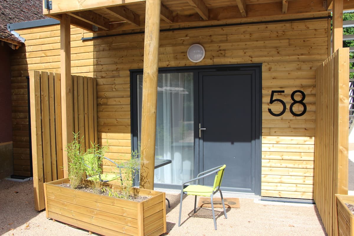 Un bâtiment en bois avec le numéro « 58 » sur la porte, un jardinier en bois contenant des plantes et une chaise verte à l’extérieur.