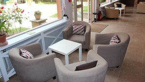 Un área de estar interior iluminada por el sol, con sillones color beige y una mesa blanca, con ventanas amplias que ofrecen vistas al exterior.